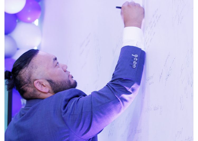 A man in a blue suit writes on a large white wall filled with signatures. Purple and white balloons are visible in the background, and the MDM Ankole Pinnada logo appears in the top right corner.