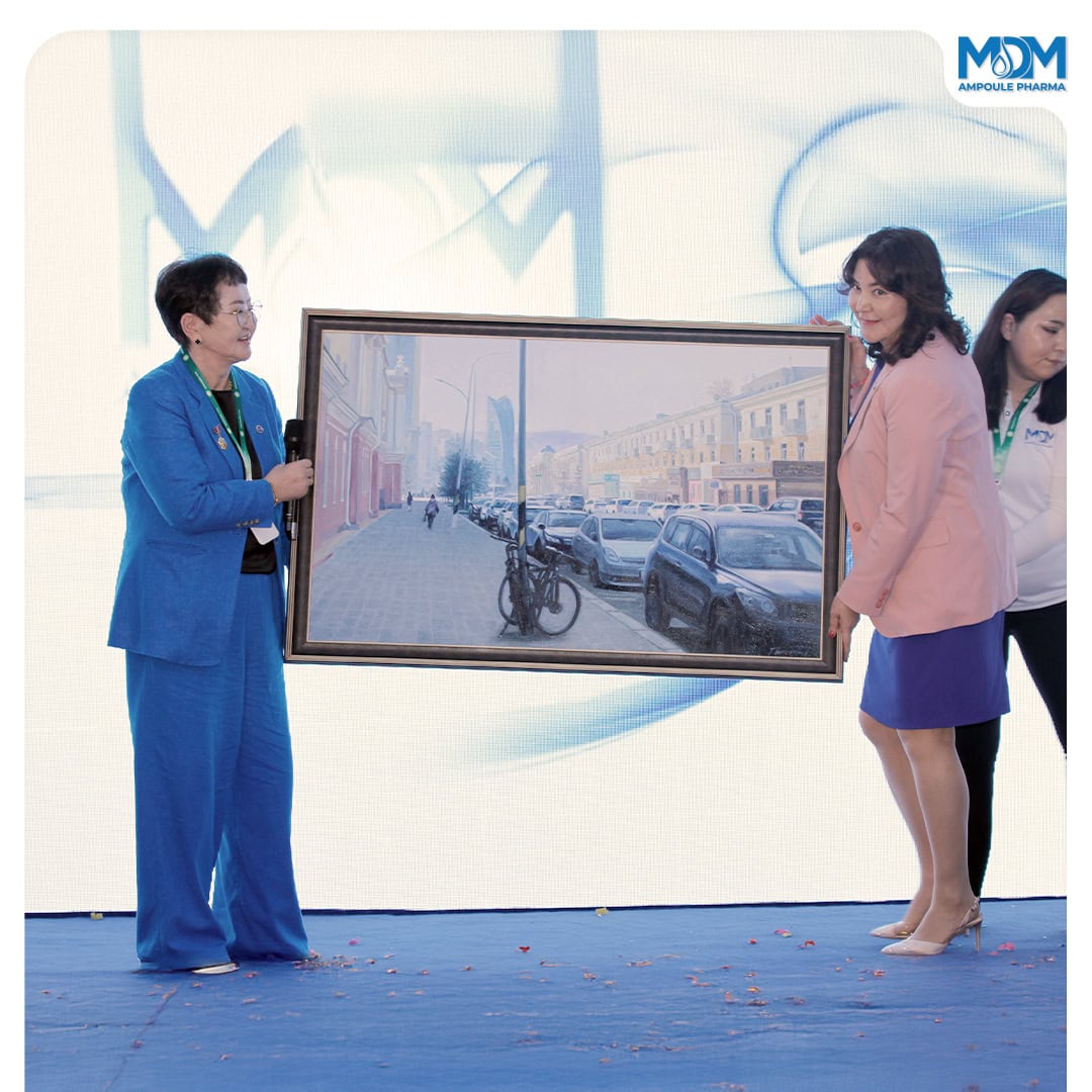 Two women stand on a stage, smiling and holding a framed painting of a city street with parked cars and bicycles. Another woman stands behind them. The backdrop displays a blue and white design with the "MDM" logo.