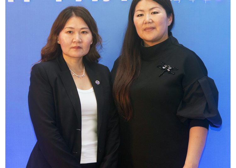 Two women pose in front of a blue background with white text. One wears a black blazer over a white top, the other wears a black dress with decorative details. Both have long, dark hair and stand side by side, looking at the camera.