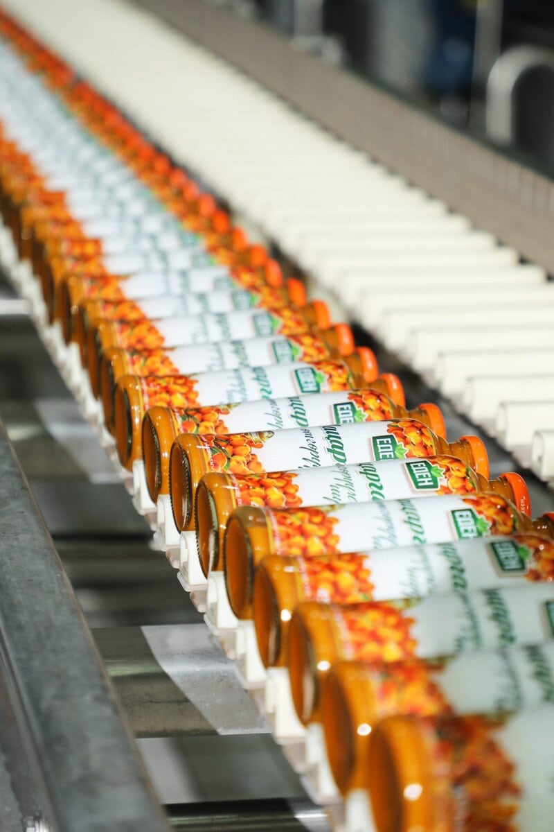 Rows of identical canned goods with orange, white, and green labels move along an automated conveyor belt in a food processing or packaging facility.