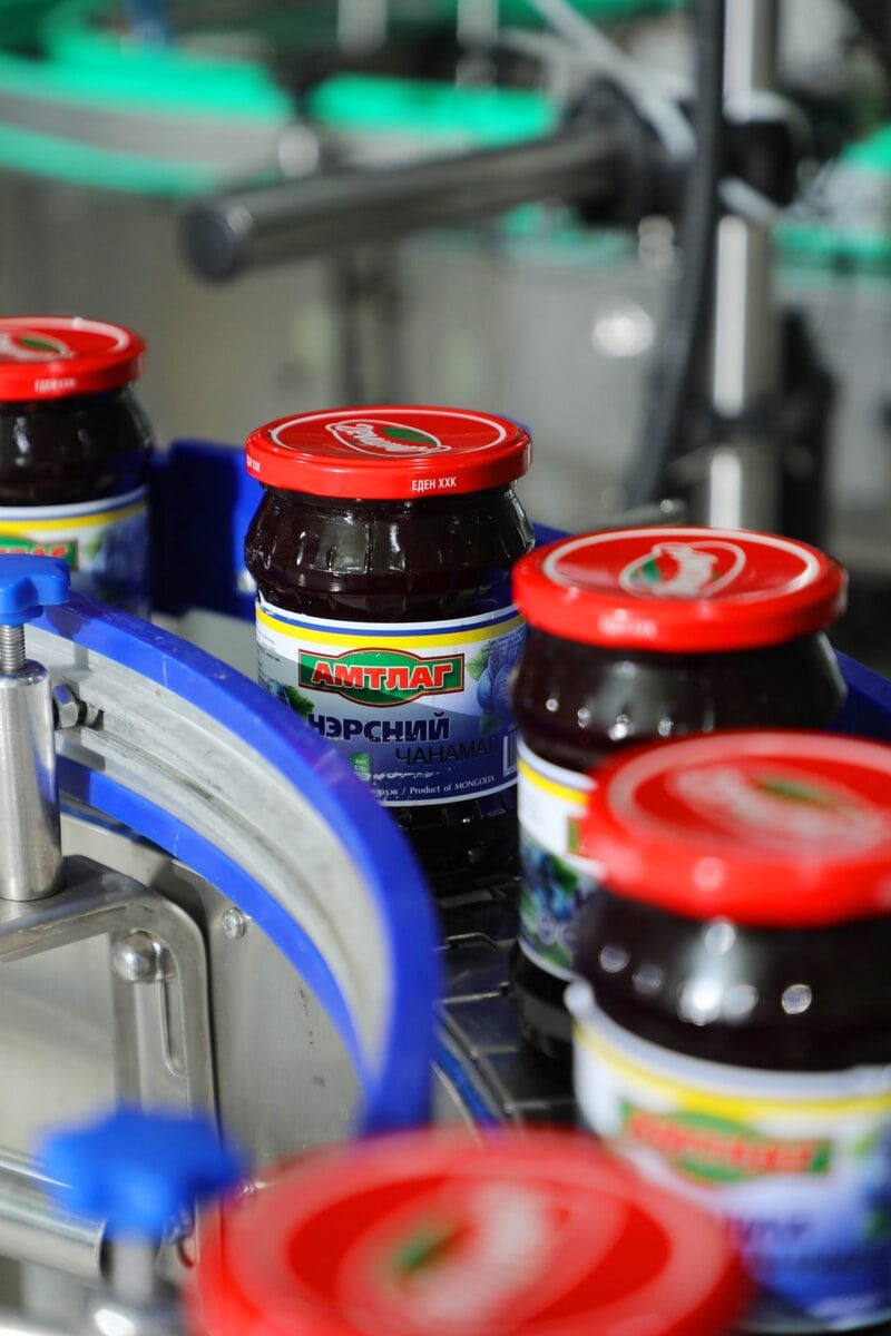 Jars of jam with red lids move along a conveyor belt in a factory setting. The labels feature text in Cyrillic and display images of berries. The scene suggests an automated food production process.