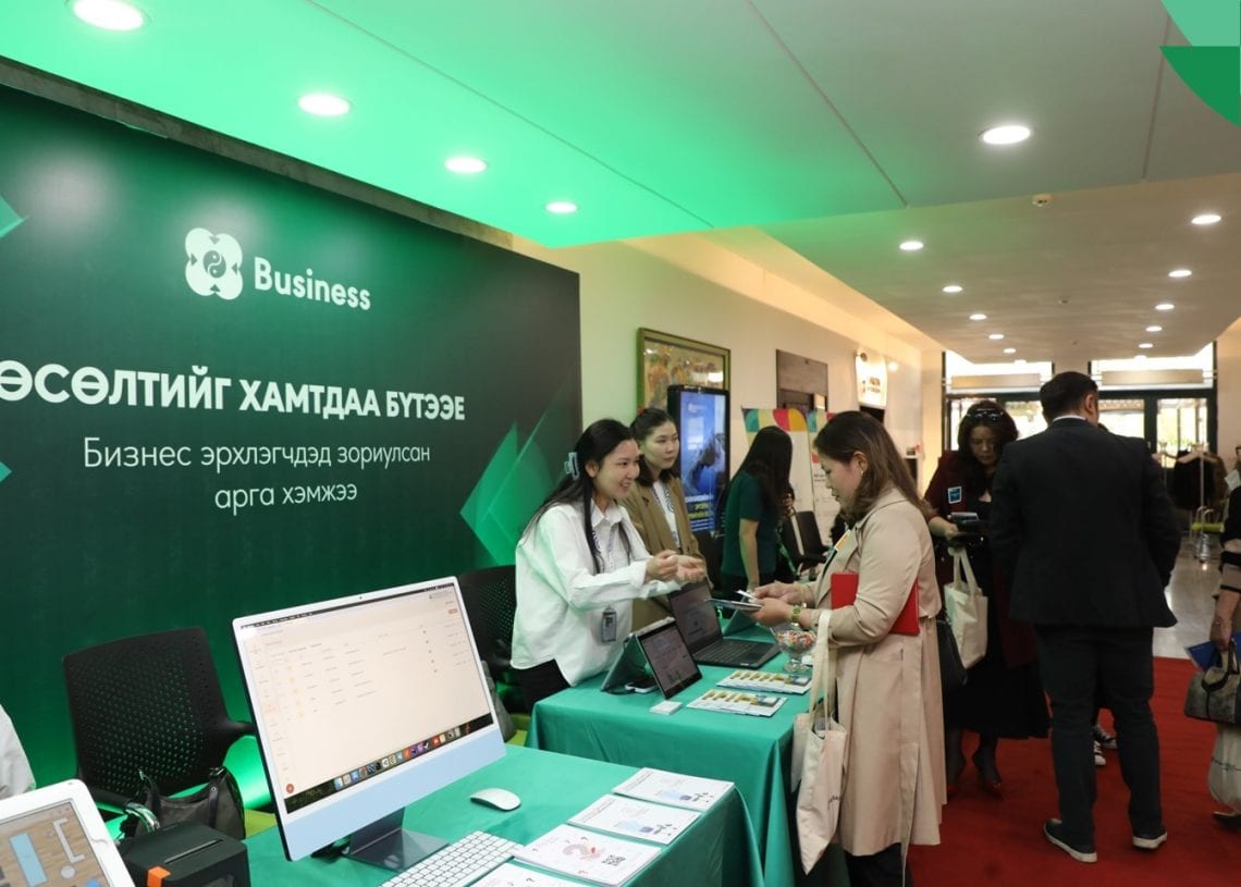 People interact at a business event booth with laptops and informational materials on tables; a green banner with Mongolian text and a "Business" icon is displayed at the back. The setting appears professional and busy.