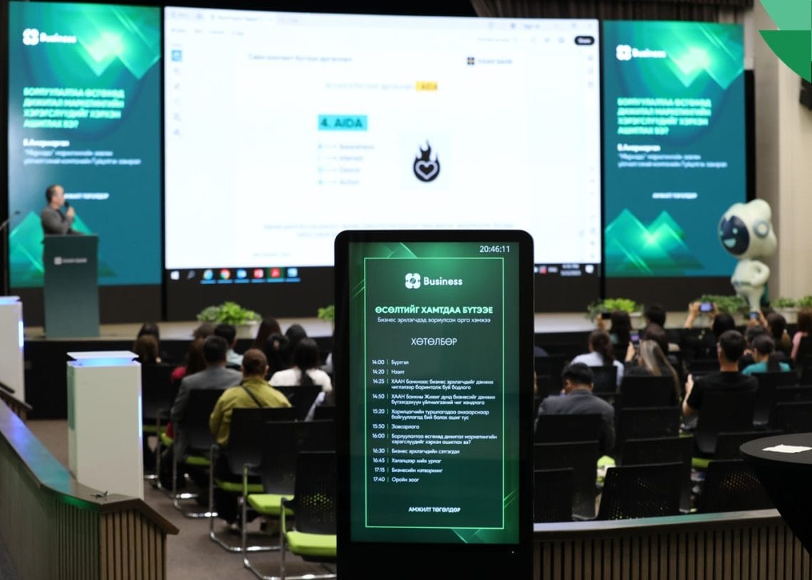 A business seminar with attendees seated in an auditorium, a speaker at the podium, presentation slides on large screens, and a digital sign showing seminar details in a modern, well-lit venue.