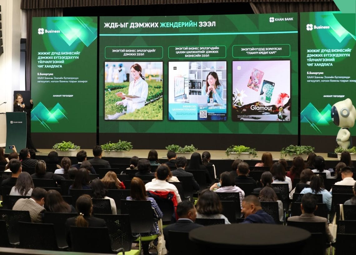 An audience sits facing a stage with large screens displaying business presentations and images of entrepreneurs. The event appears professional, with a presenter at a podium and plants decorating the stage.