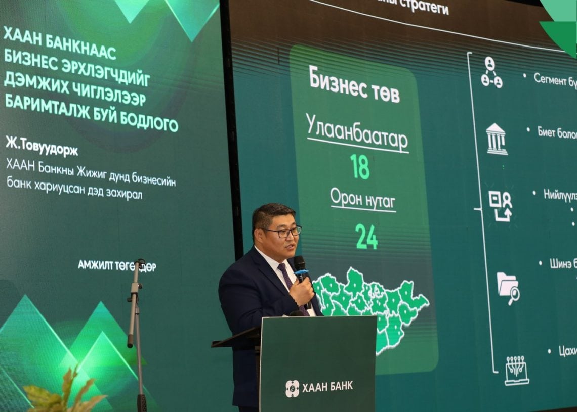 A man in a dark suit and glasses speaks at a podium with the Khaan Bank logo. Behind him is a large screen showing business statistics and a map of Mongolia, with text in Mongolian.