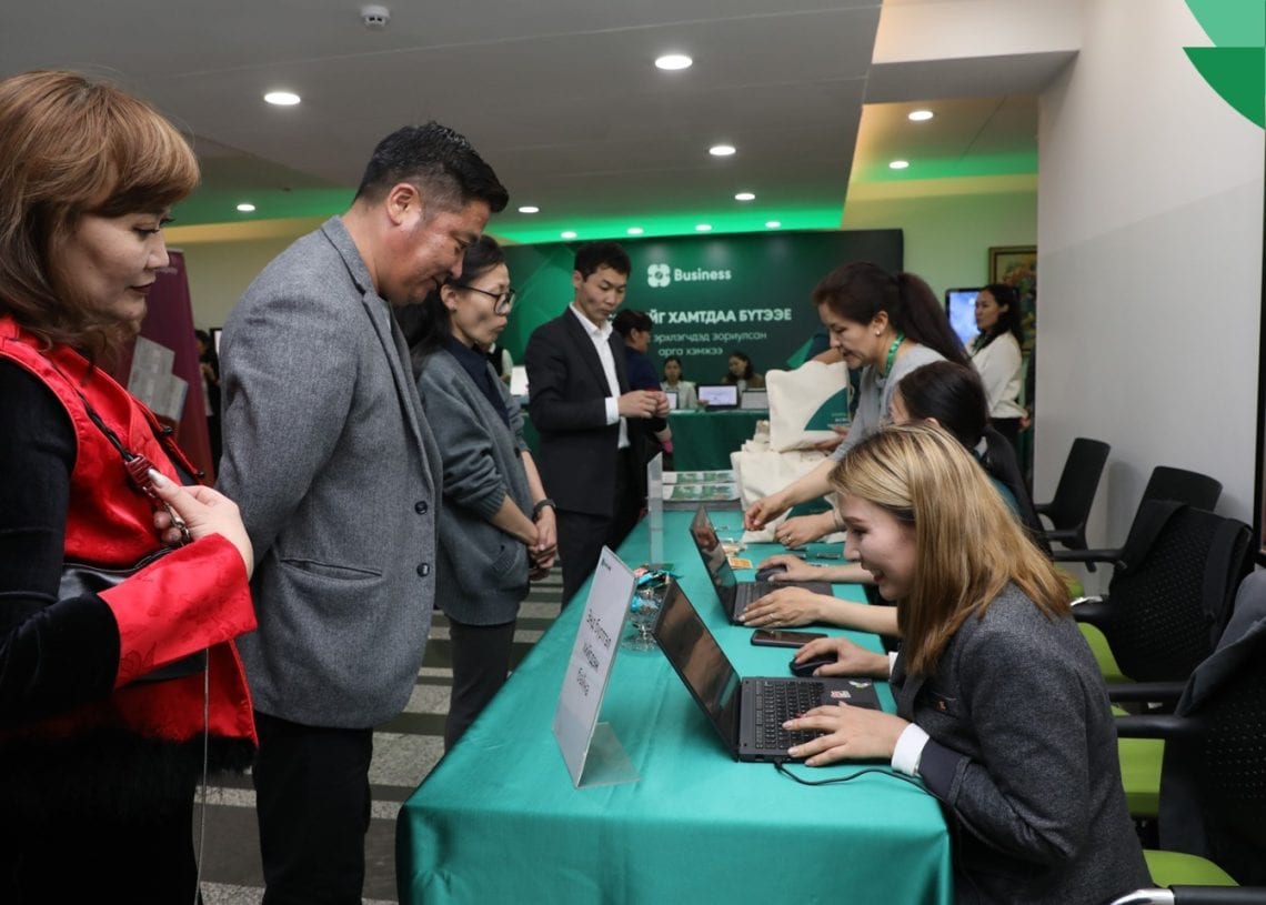 People are signing in at a registration desk covered with a green tablecloth. Several staff members are assisting visitors, and informational signs are displayed on the table. The setting appears to be an event or conference.
