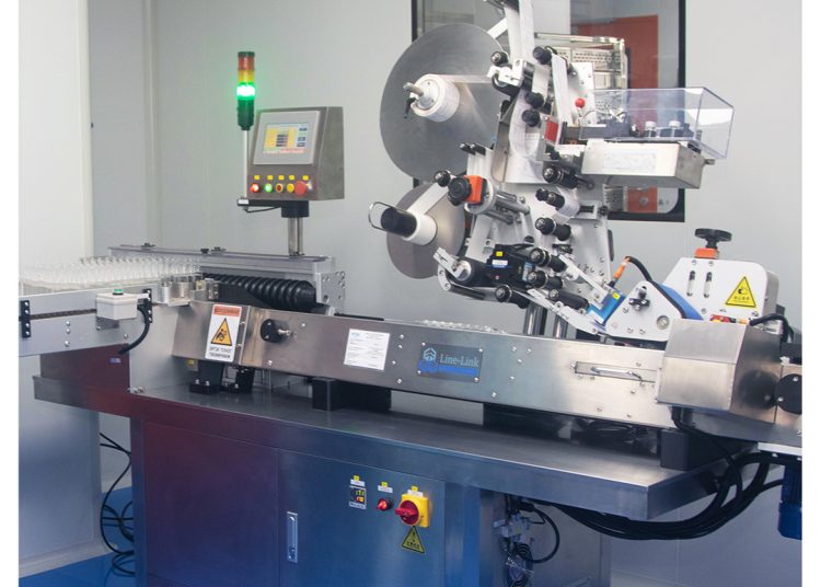 A pharmaceutical labeling machine in a clean, brightly lit lab, with a control panel, roll of labels, warning signs, and transparent safety covers. The MDM Ampoule Pinhas logo is visible in the top right corner.