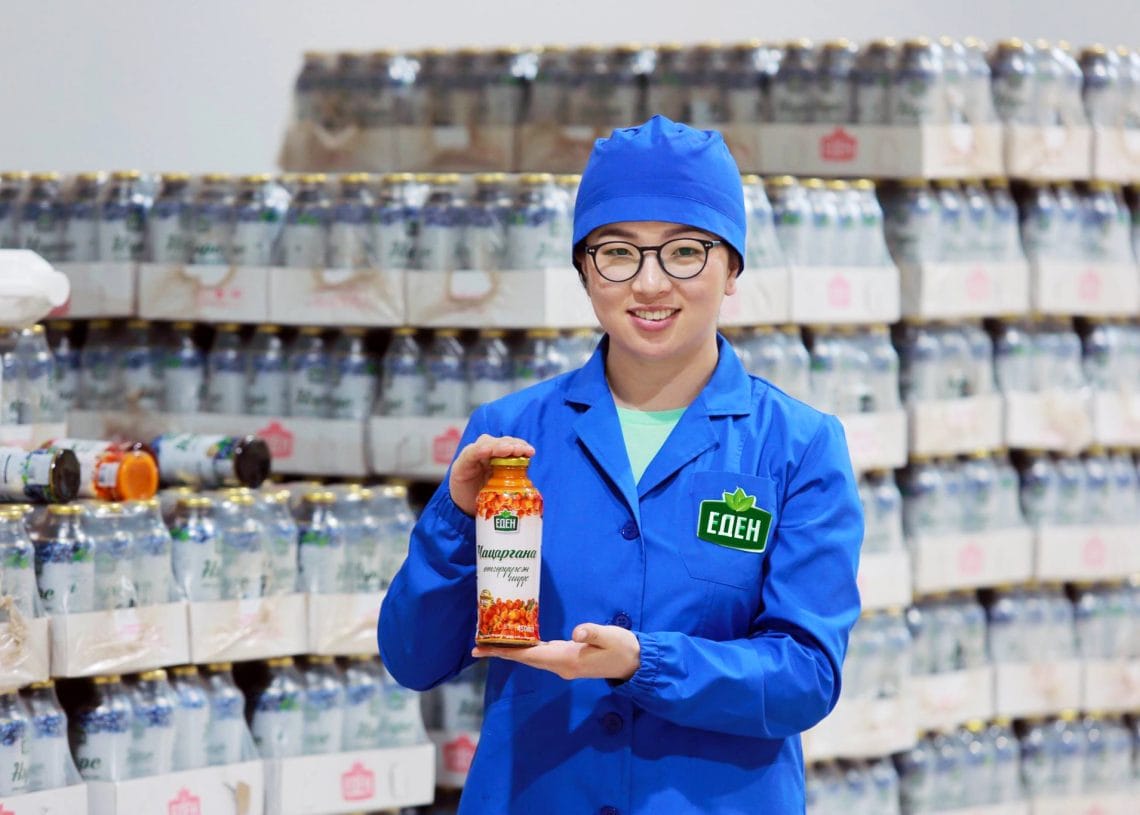 A person wearing a blue lab coat and cap stands in front of stacked jars and smiles while holding up a jar labeled "VEGGIPAN," displaying it to the camera.