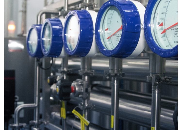 A row of industrial pressure gauges with blue rims mounted on metal pipes in a factory setting. The background shows more pipes and valves. Logo at the top right reads "MDM ANGOULÊ PHARMA.