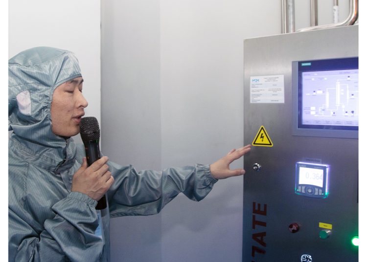 A person in a blue protective suit speaks into a microphone while pointing at a control panel with a digital display and various buttons in a clean, industrial environment.