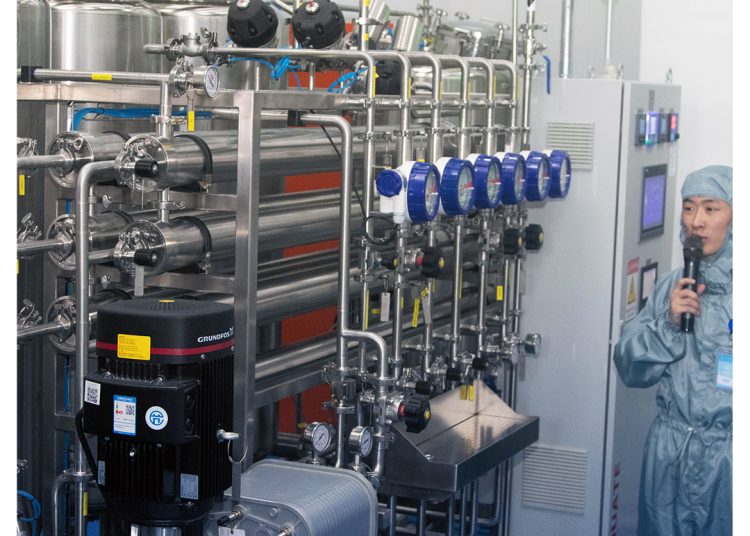 A person in protective clothing speaks into a microphone beside an industrial machine with many pipes, gauges, and control panels in a clean, modern facility.