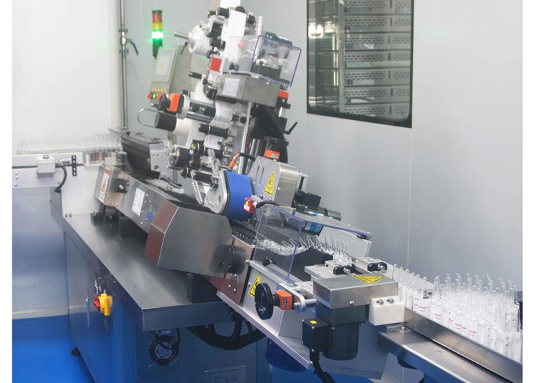 A modern pharmaceutical labeling machine in a clean, sterile environment applies labels to rows of transparent ampoules moving along a conveyor belt. The MOM Ampoule Pramina logo is visible in the top right corner.