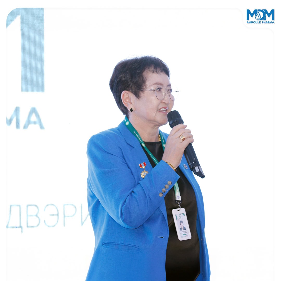 An older woman in a blue blazer speaks into a microphone at an event. She has short hair, glasses, and wears a lanyard and several pins. There is a white background with blue text and a logo in the top right corner.
