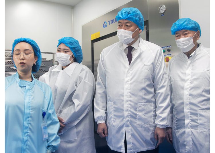Four people in lab coats, blue hairnets, and face masks stand in a laboratory with medical equipment in the background. One person appears to be explaining something to the group.