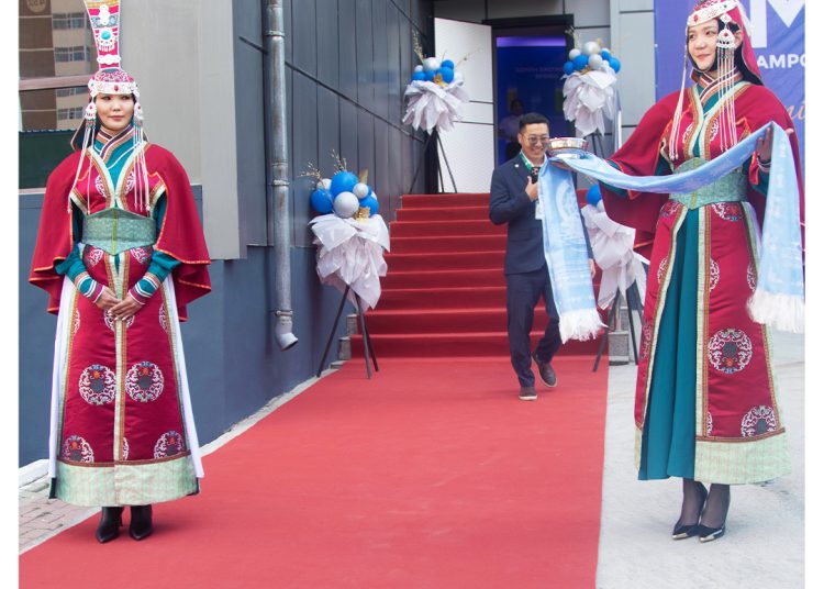 Two women in traditional Mongolian attire stand on either side of a red carpet holding ceremonial scarves; a man in a suit walks down the steps in the background near blue and white balloons.