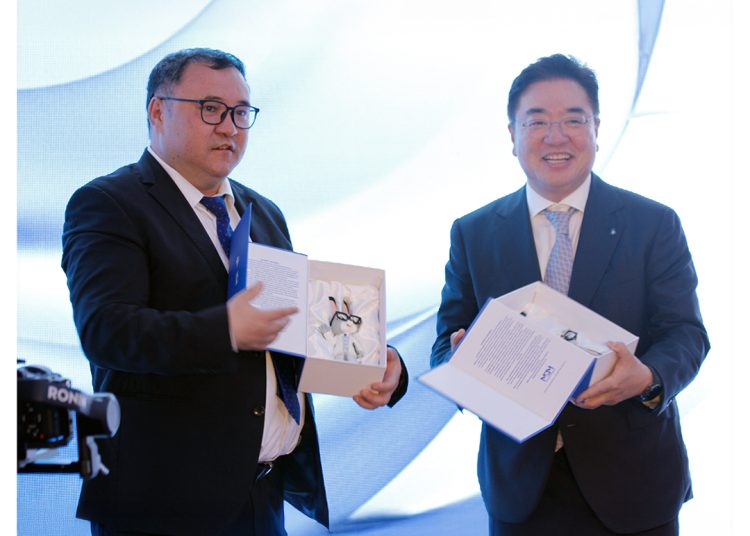 Two men in suits stand side by side at an event, each holding an open box with a product inside. Both are smiling. The background features a light blue abstract design and an MDM Ampoule Phanna logo in the top right corner.
