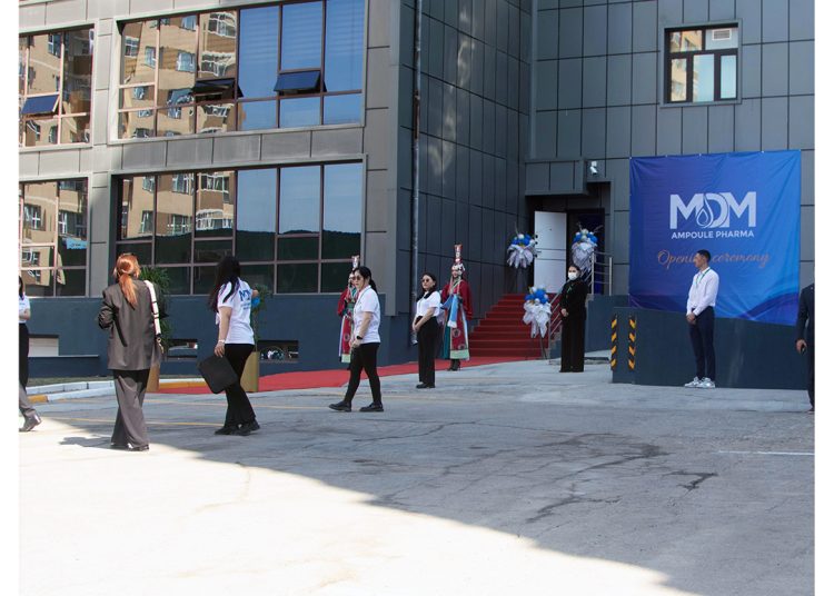 People walk near the entrance of a modern building decorated with blue and white balloons for an opening ceremony. A large blue banner with "Open Ceremony" and the MOM Ampoule Pharma logo is visible.
