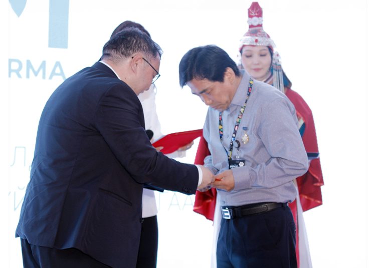 Two men standing on stage, one presenting an award or certificate to the other. A woman in traditional attire stands in the background, observing the ceremony.
