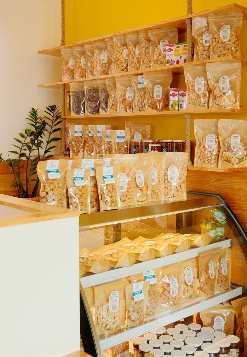 Shelves and a display case in a store filled with many clear bags of snacks, jars, and containers. The background wall is yellow, and there is a green plant in the corner.