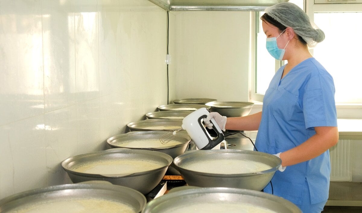 A person in scrubs, a hairnet, gloves, and a face mask uses a hand mixer to blend ingredients in large metal bowls lined up on a counter in a clean, industrial kitchen setting.