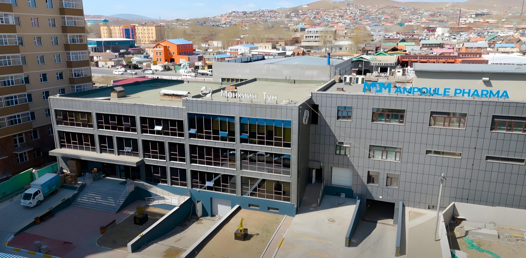 A modern gray building labeled "Ampoule Pharma" stands next to a multi-story building in an urban area, with a mix of colorful houses and hills visible in the background under a clear sky.