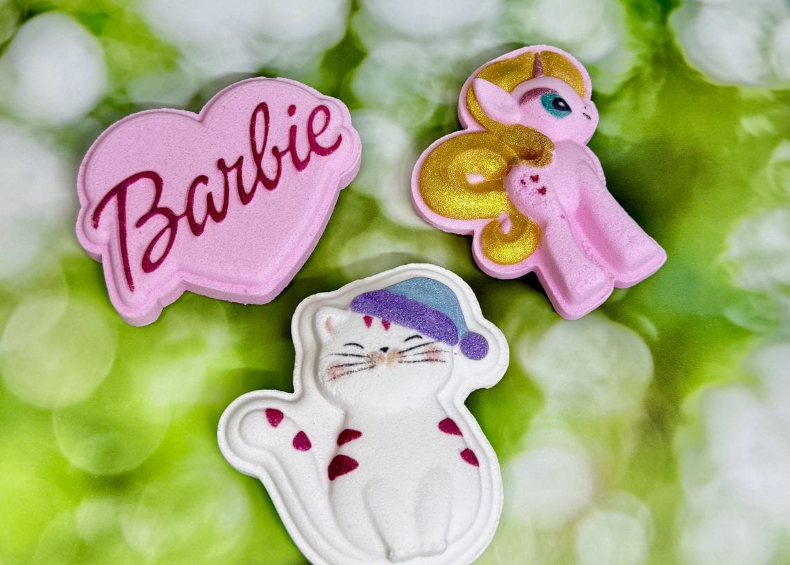 Three bath bombs on a green and white blurred background: a pink heart-shaped one with "Barbie" written in cursive, a pink and gold unicorn, and a white cat wearing a purple sleeping cap.