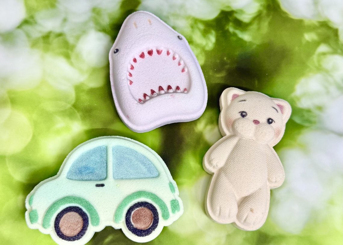 Three pastel-colored cookies shaped like a green car, a smiling beige cat, and a white shark with an open mouth, are arranged on a blurred green and white background.