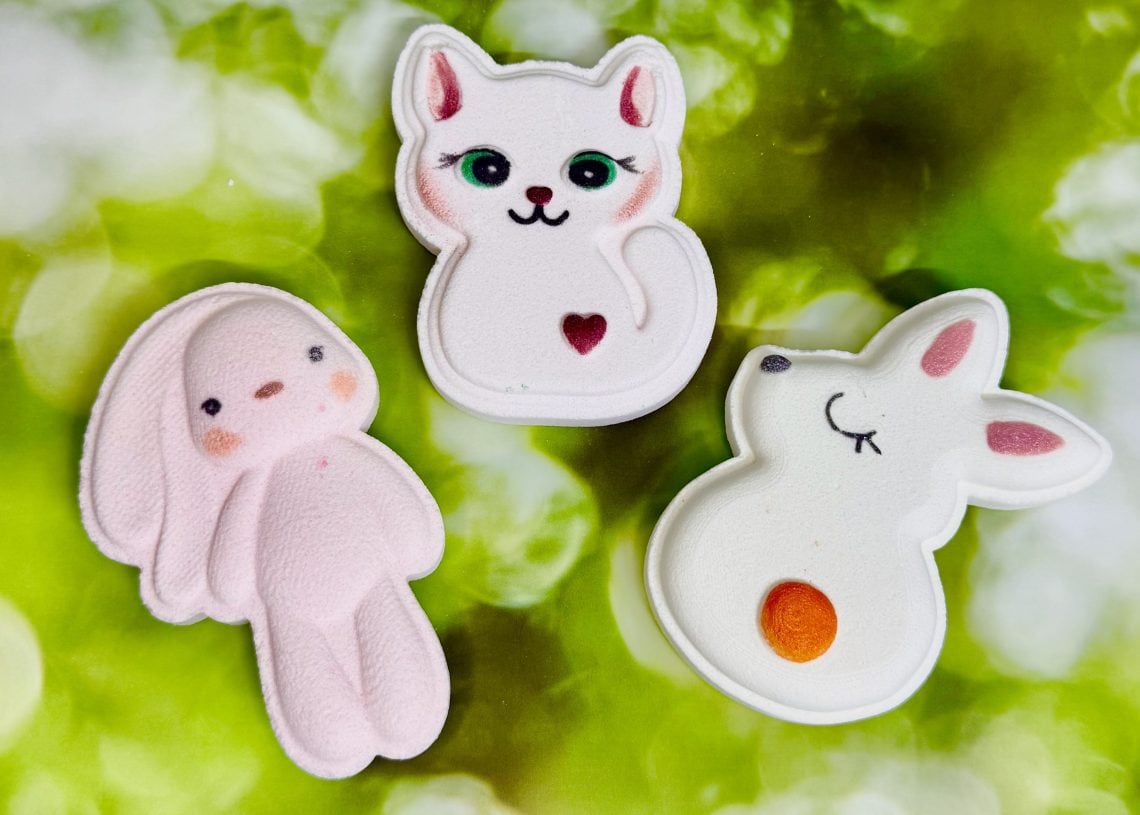 Three pastel plush toys—a pink bunny, a white cat with green eyes and a heart mark, and a white mouse with an orange spot—are arranged on a green, nature-inspired background.