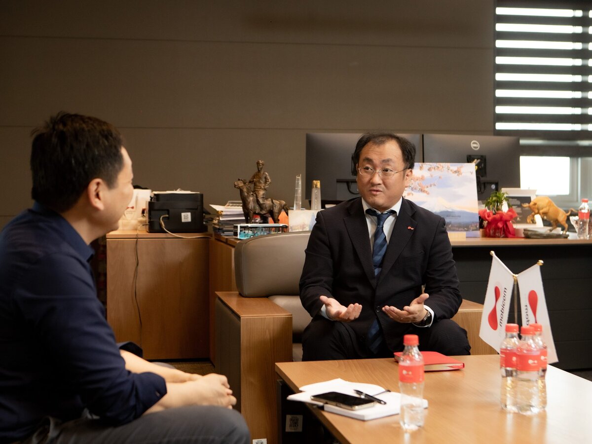 Two men sit across from each other in an office, engaged in conversation. One man gestures with his hands while the other listens. A desk with documents, two water bottles, and flags is in the foreground.