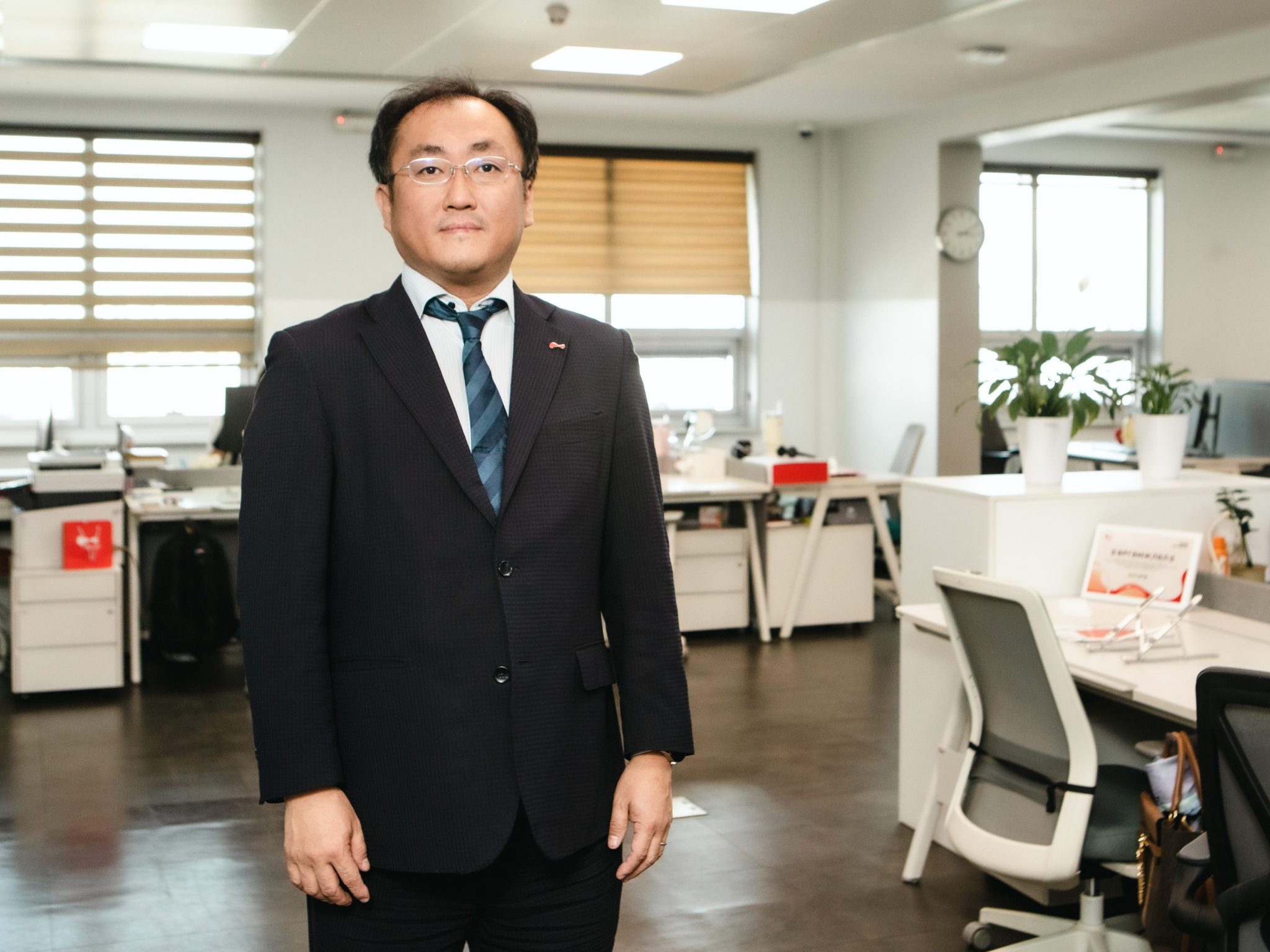 A man in a dark suit and tie stands in a modern office space with desks, chairs, computers, and potted plants visible in the background.
