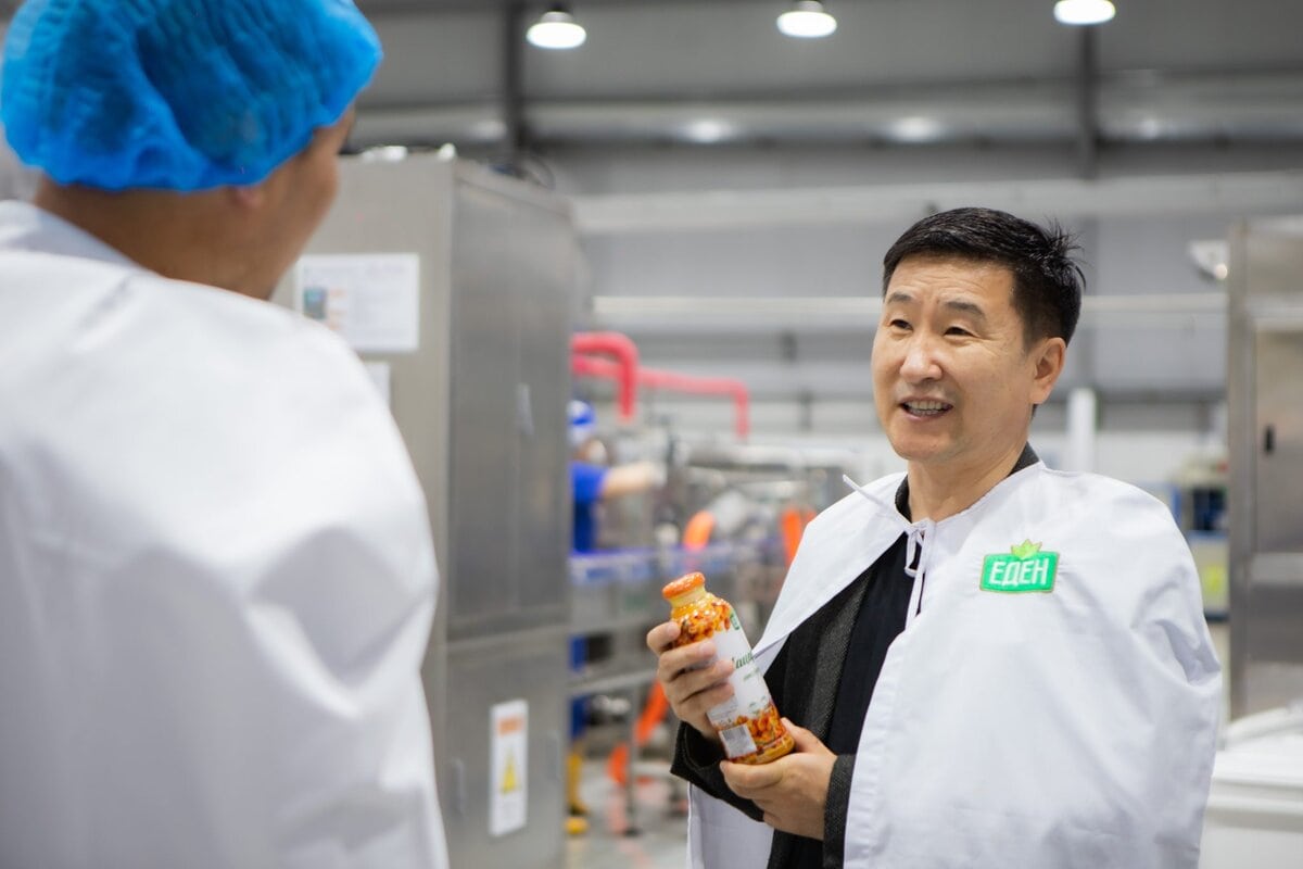 A man in a white lab coat holding a jar of food speaks to another person in a lab coat and blue hairnet inside a food processing facility. Industrial equipment is visible in the background.