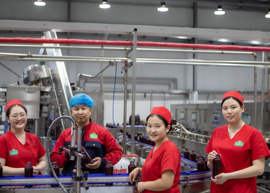 Four factory workers, three women and one man, stand by a conveyor belt with glass bottles in a modern production facility. The workers wear red uniforms and the man wears a blue hair net; all are smiling at the camera.