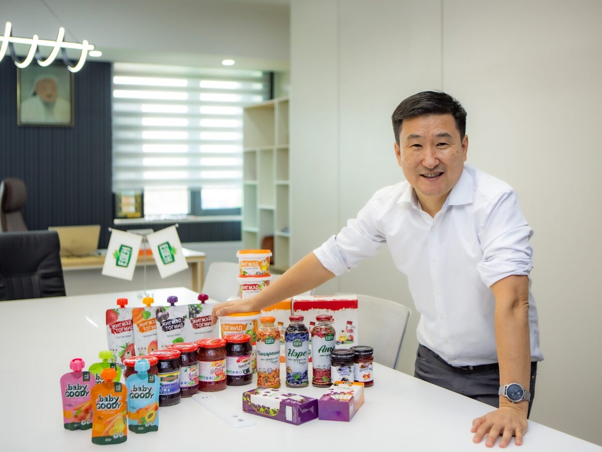 A man in a white shirt smiles while standing next to a table displaying various colorful food and drink products in a modern office setting.