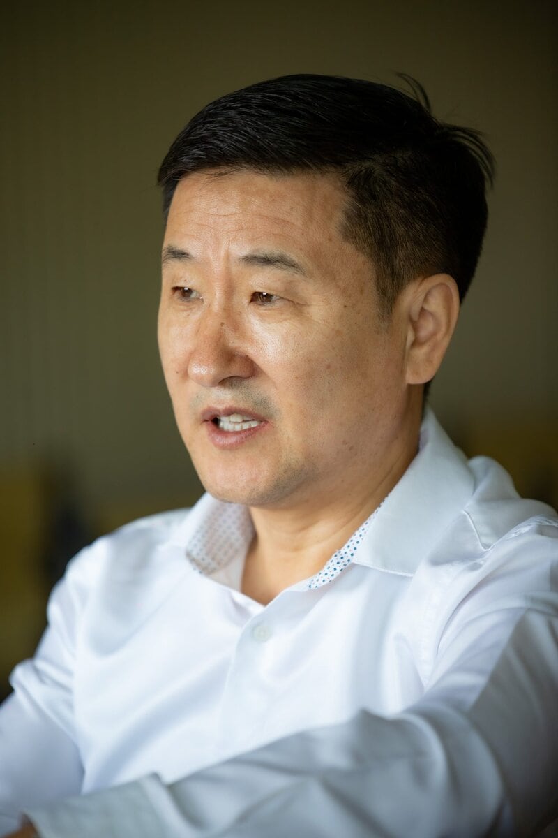 A man with short black hair wearing a white collared shirt is speaking or engaged in conversation, with a neutral indoor background.