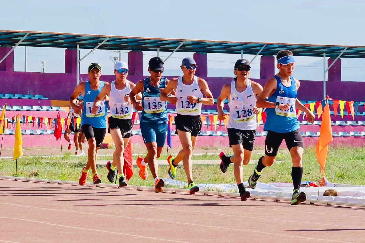 Seven male athletes run on a track during a race, wearing numbered bibs, sunglasses, and athletic gear. Colorful flags and empty stadium seats are visible in the background. The weather appears sunny and clear.