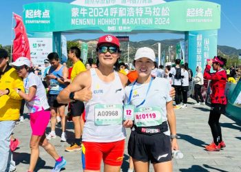 Two marathon runners pose and smile at the start line of the Mengniu Hohhot Marathon 2024, wearing race bibs and athletic gear, with the event banner and other participants in the background.