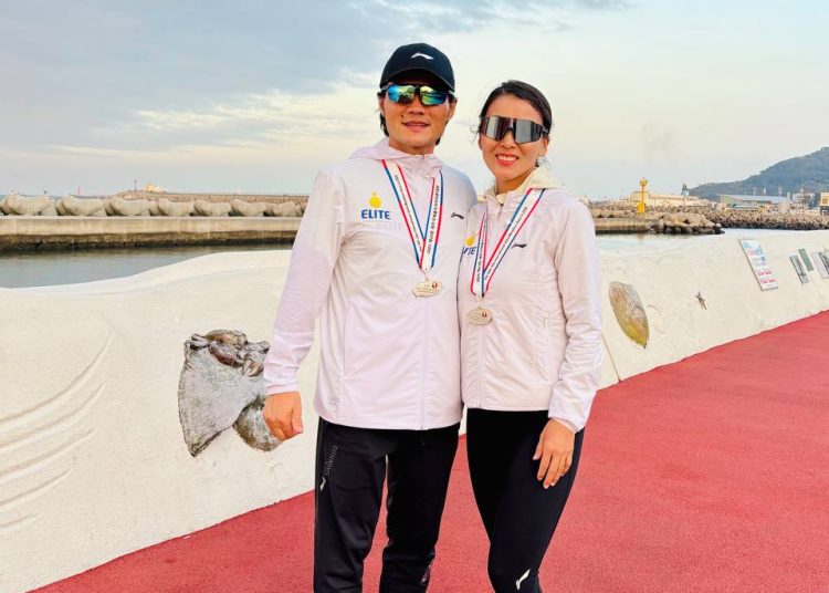 Two runners wearing white jackets, black pants, sunglasses, and medals stand smiling on a red track by a seaside promenade, with cloudy sky and distant mountains in the background.