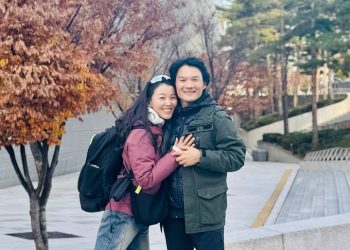 A smiling couple embraces outdoors on a paved walkway lined with autumn trees. The woman wears a pink jacket and lifts one leg playfully, while the man wears a green jacket. Both have backpacks.