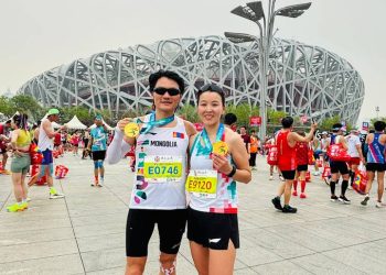 Two marathon runners pose smiling with medals in front of the Beijing National Stadium, also known as the Bird's Nest. They wear race bibs and sportswear, surrounded by other participants and spectators.