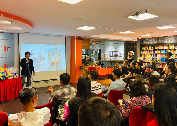 A man gives a presentation to an attentive audience in a bookstore, with a projected image on the screen behind him and colorful items on a table beside him. Shelves of books and wall art are visible in the background.