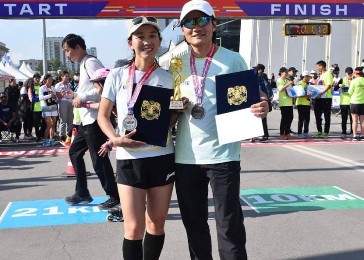 Two runners stand smiling at the finish line of the Ulaanbaatar International Marathon 2024, holding medals, certificates, and a trophy, with the marathon banner visible above them. Other participants are in the background.