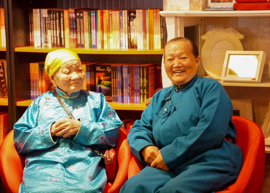 Two elderly women wearing traditional blue clothing sit and smile in a brightly lit room with bookshelves and framed photos in the background. One woman wears a yellow headscarf, the other has short hair.