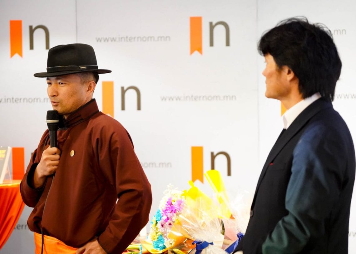 Two men stand in front of a backdrop with "internom.mn" and orange logos. One man, in a traditional outfit and black hat, speaks into a microphone; the other, in a suit, looks on. There are gift bouquets on a table nearby.