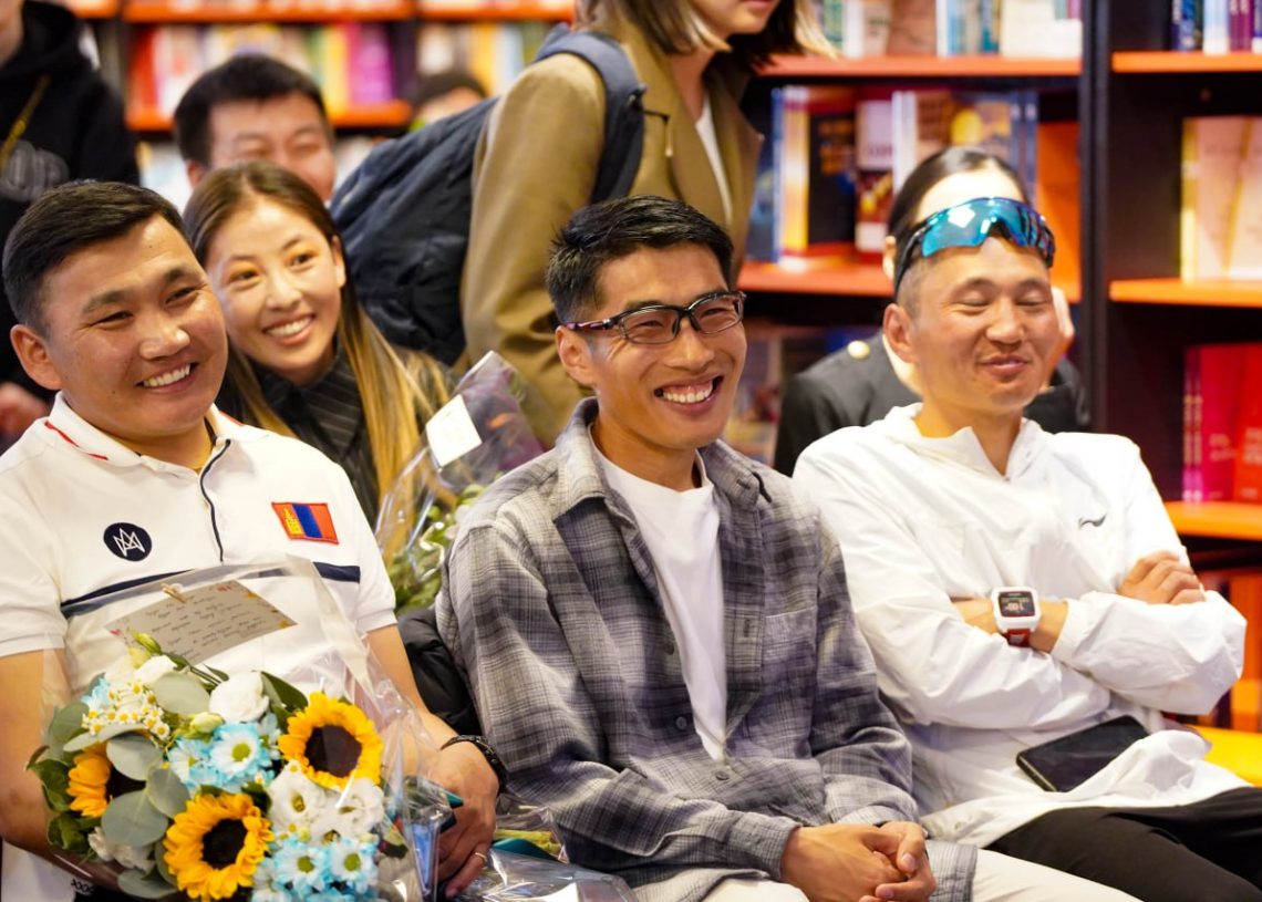 A group of smiling people sit closely together in a brightly lit bookstore. One person holds a bouquet of flowers, and shelves filled with colorful books are visible in the background.