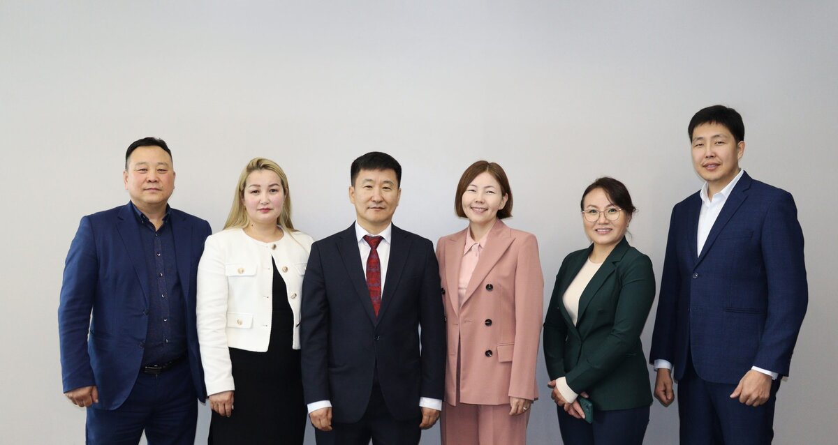 Six people in business attire stand in a row against a plain light gray background, smiling at the camera. The group includes three men in suits and three women in professional outfits.