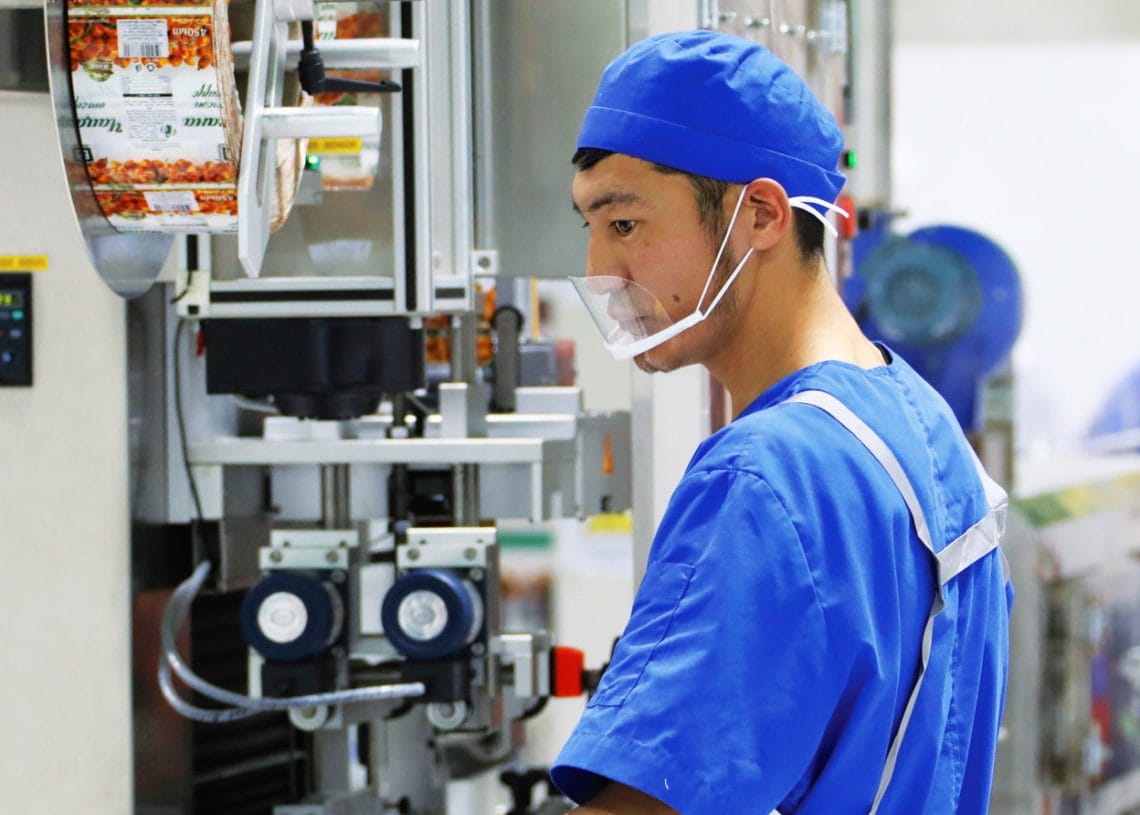 A factory worker in blue uniform and cap operates food packaging machinery, surrounded by rolls of printed packaging film. He wears a face shield, gloves, and a white apron inside a clean, industrial facility.