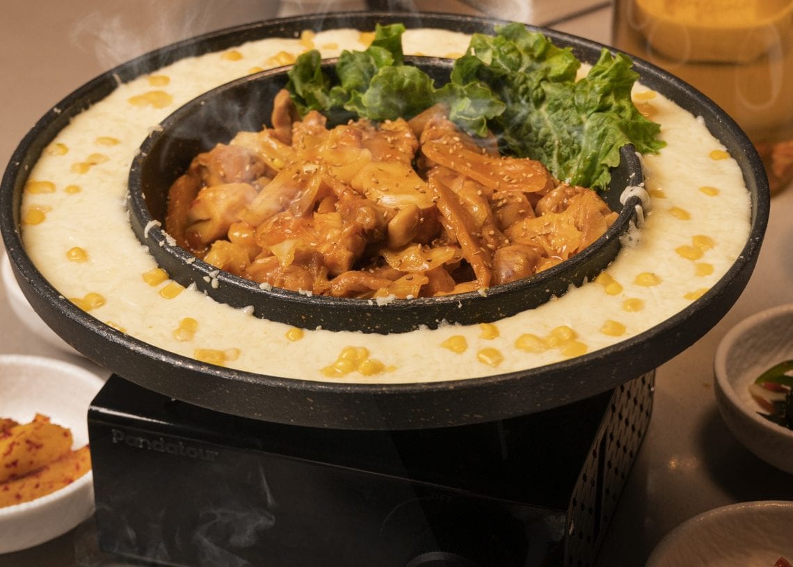 A sizzling hot pot filled with marinated meat and greens sits atop a stove, surrounded by a ring of melted cheese and corn. Side dishes and a drink are on the table, creating a cozy dining atmosphere.