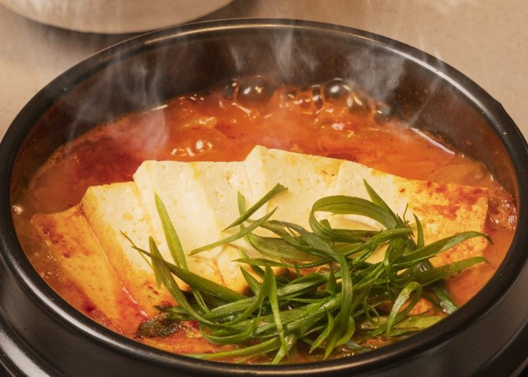 A steaming bowl of Korean tofu stew topped with fresh green herbs sits on a table, with a side dish of pickled radish in the background.