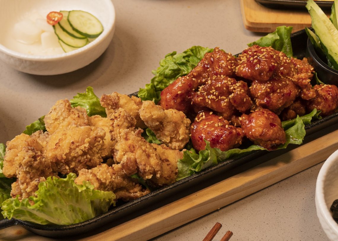 A platter with two types of fried chicken: golden crispy pieces and glazed spicy pieces, garnished with sesame seeds and served on leafy greens, surrounded by small side dishes.