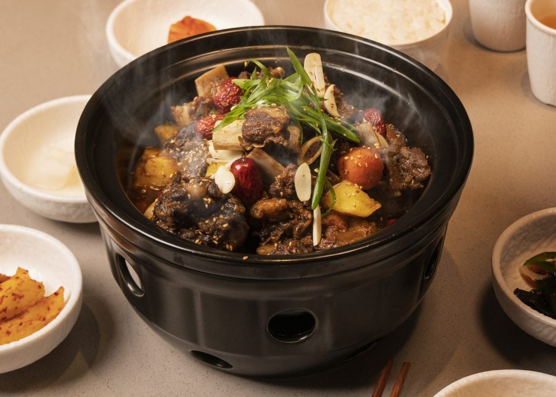 A steaming hot pot filled with braised meat, vegetables, and herbs sits on a table surrounded by bowls of rice, kimchi, tea, and various side dishes. Chopsticks rest nearby.